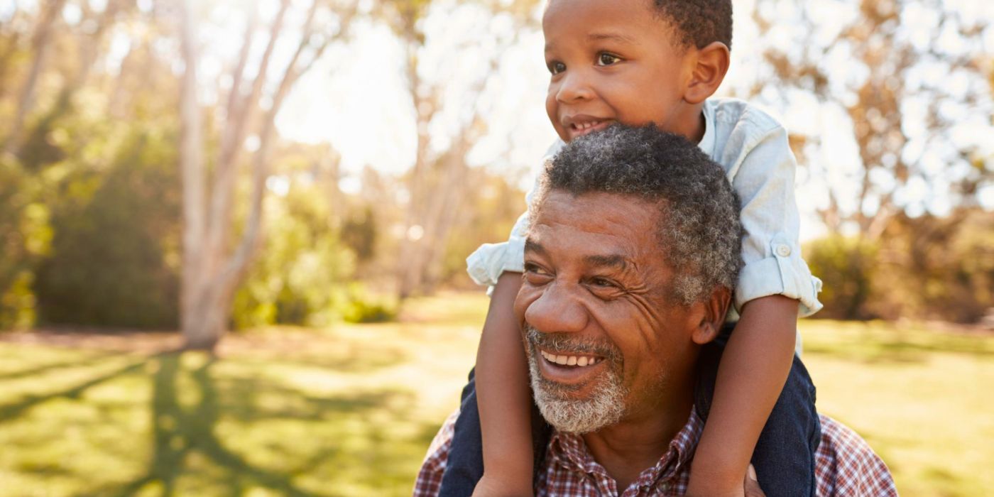 Smiling older man gives a young boy a piggyback ride outdoors on a sunny day in a park.