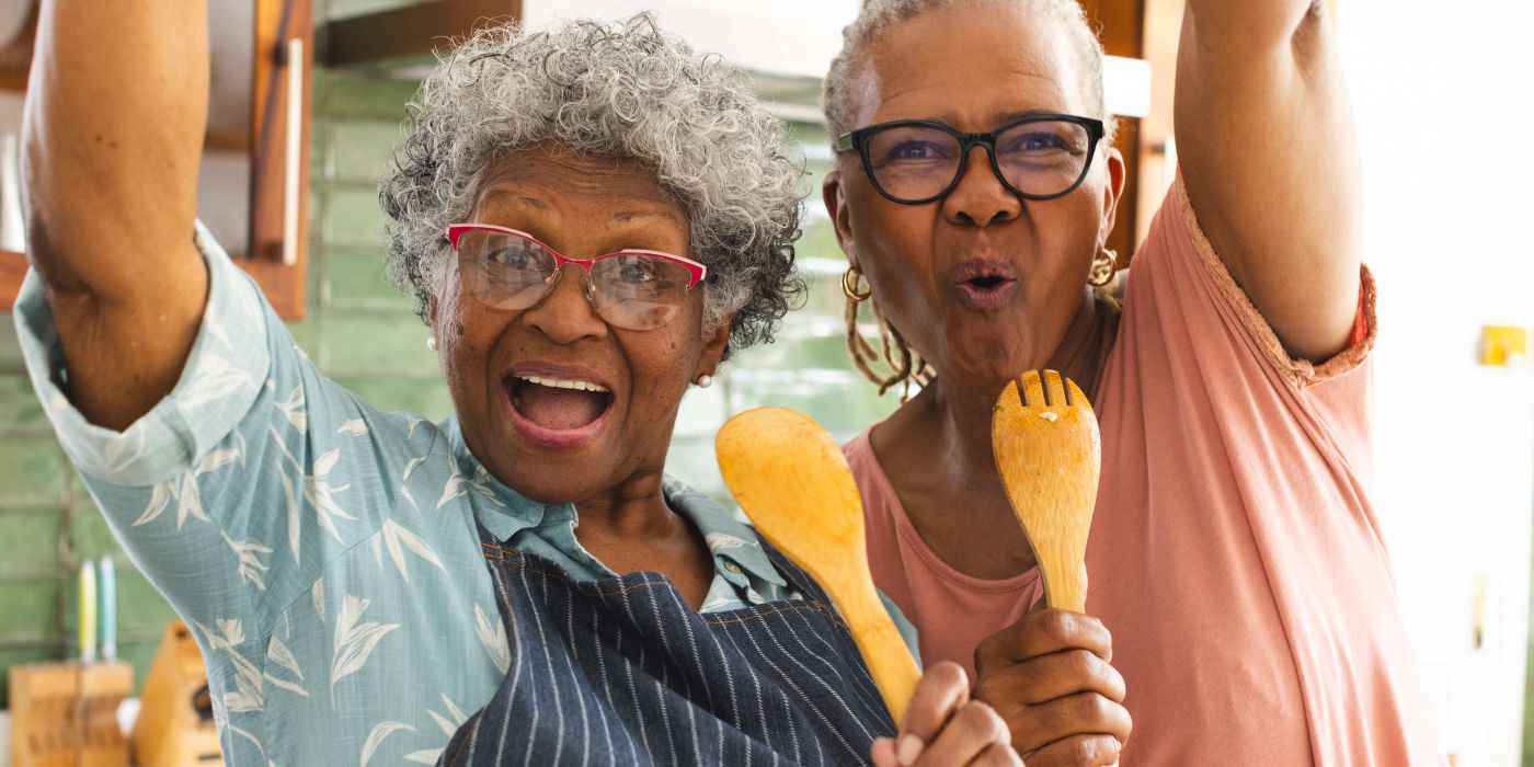 Two joyful older women in a kitchen, holding wooden utensils and smiling energetically at the camera.