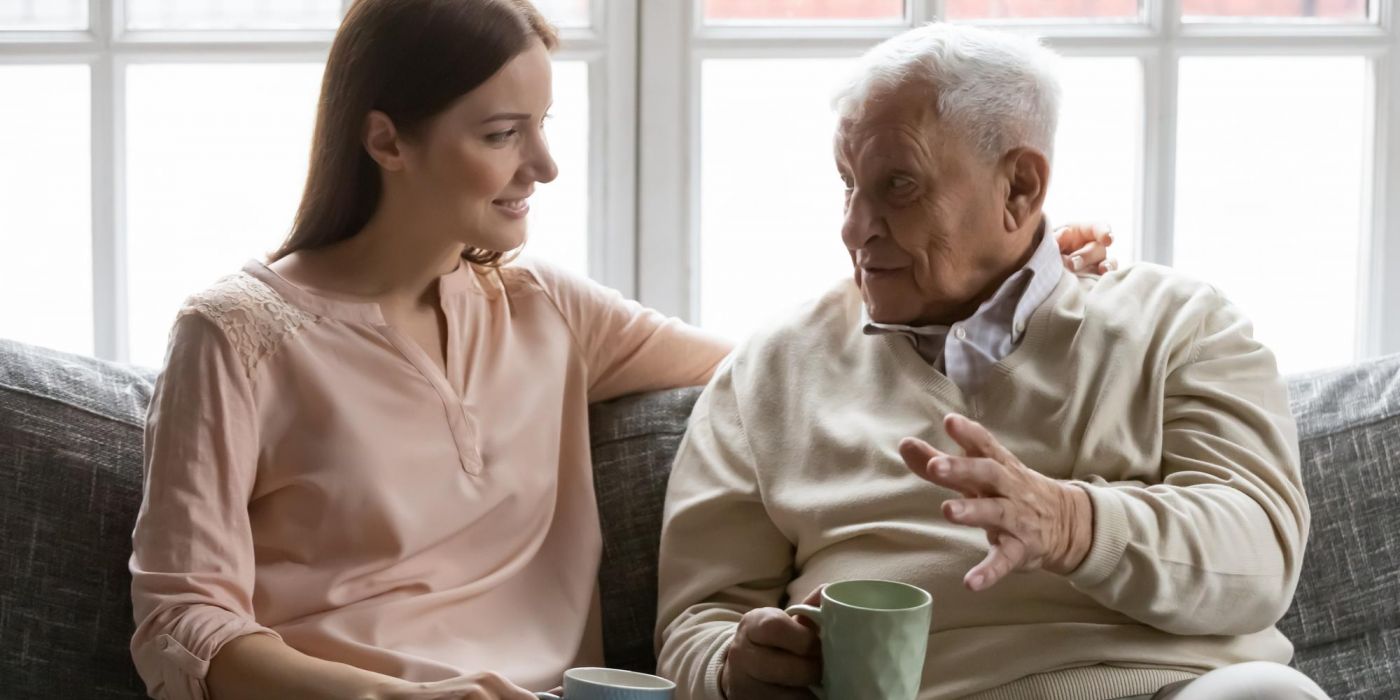 A young woman and an elderly man sitting on a sofa talking while holding mugs.