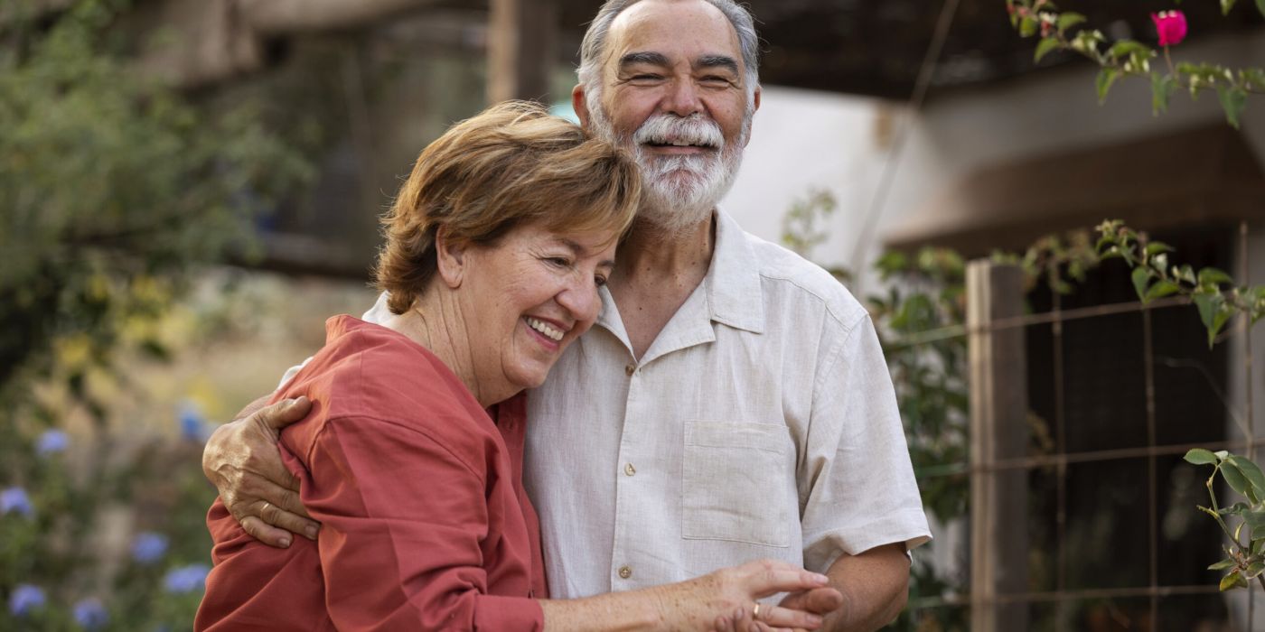 Smiling elderly couple hugging outdoors in a garden, both looking happy and relaxed.