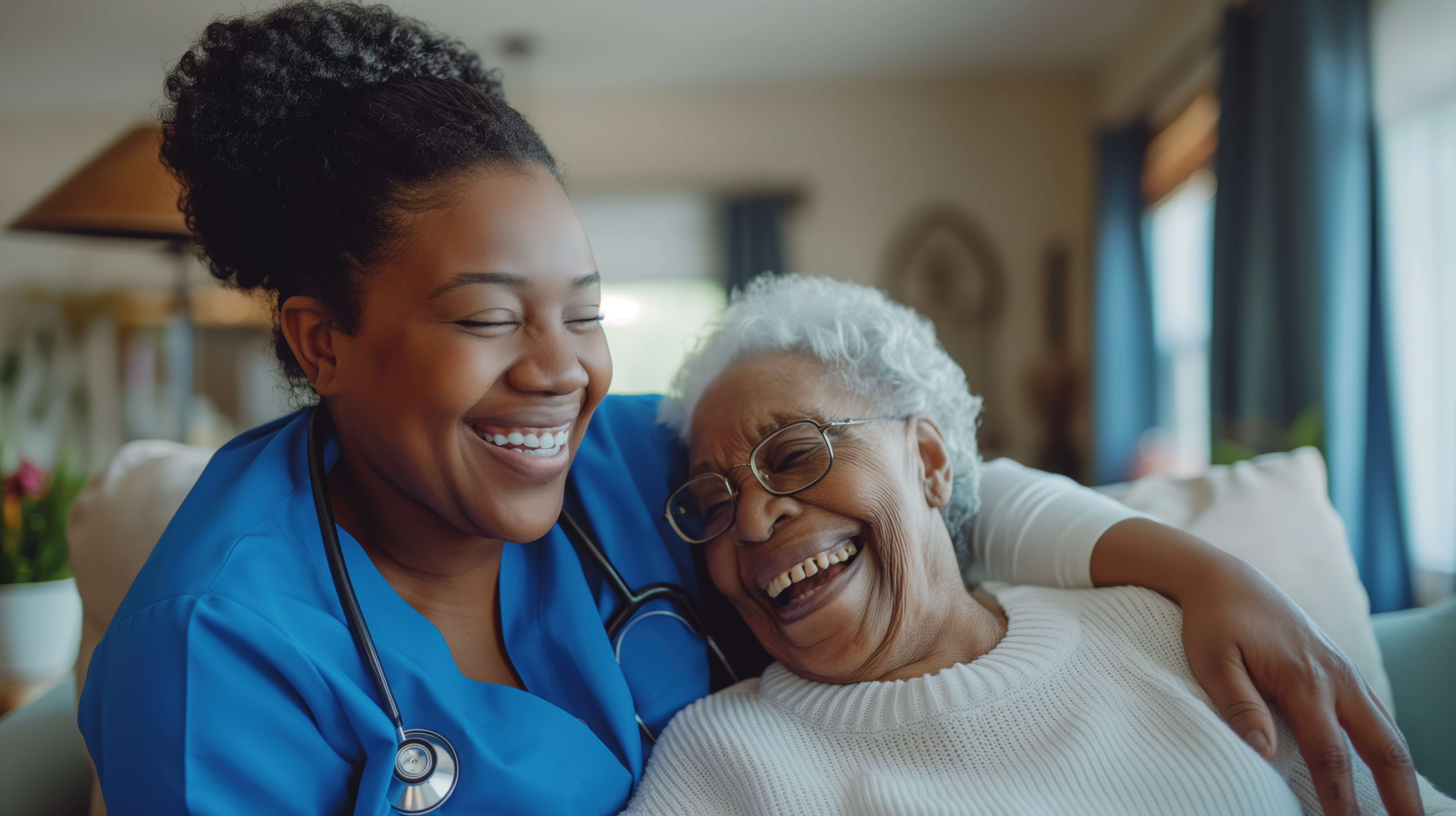 Nurse in blue scrubs with stethoscope hugs and laughs with an elderly woman wearing glasses and a white sweater.
