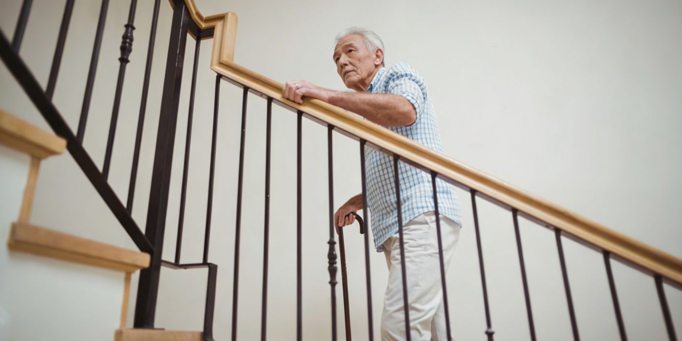 Older man in a checkered shirt holding a railing while walking up a staircase indoors.