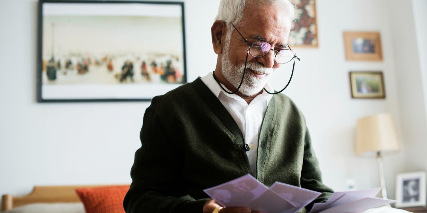 An elderly man with a white beard and glasses smiles while looking at old photographs in his bedroom.