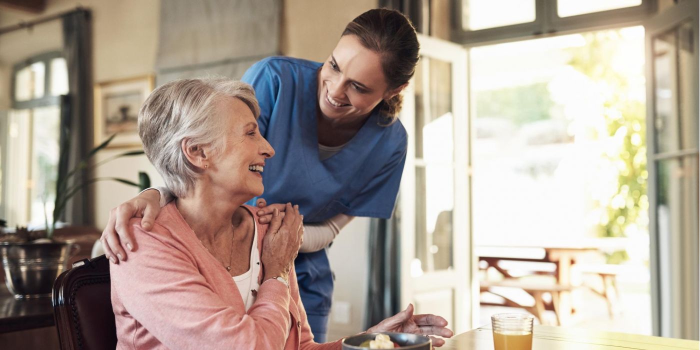 Caregiver in blue scrubs smiling at a seated elderly woman at a table with a bowl and a glass of juice.