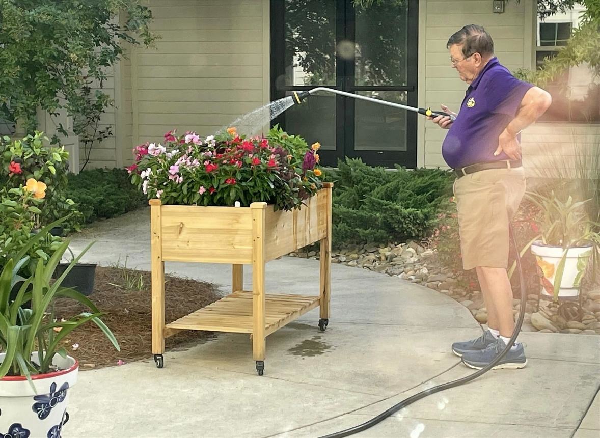 Man watering colorful flowers in a wooden planter on wheels in a garden near a house on a sunny day.