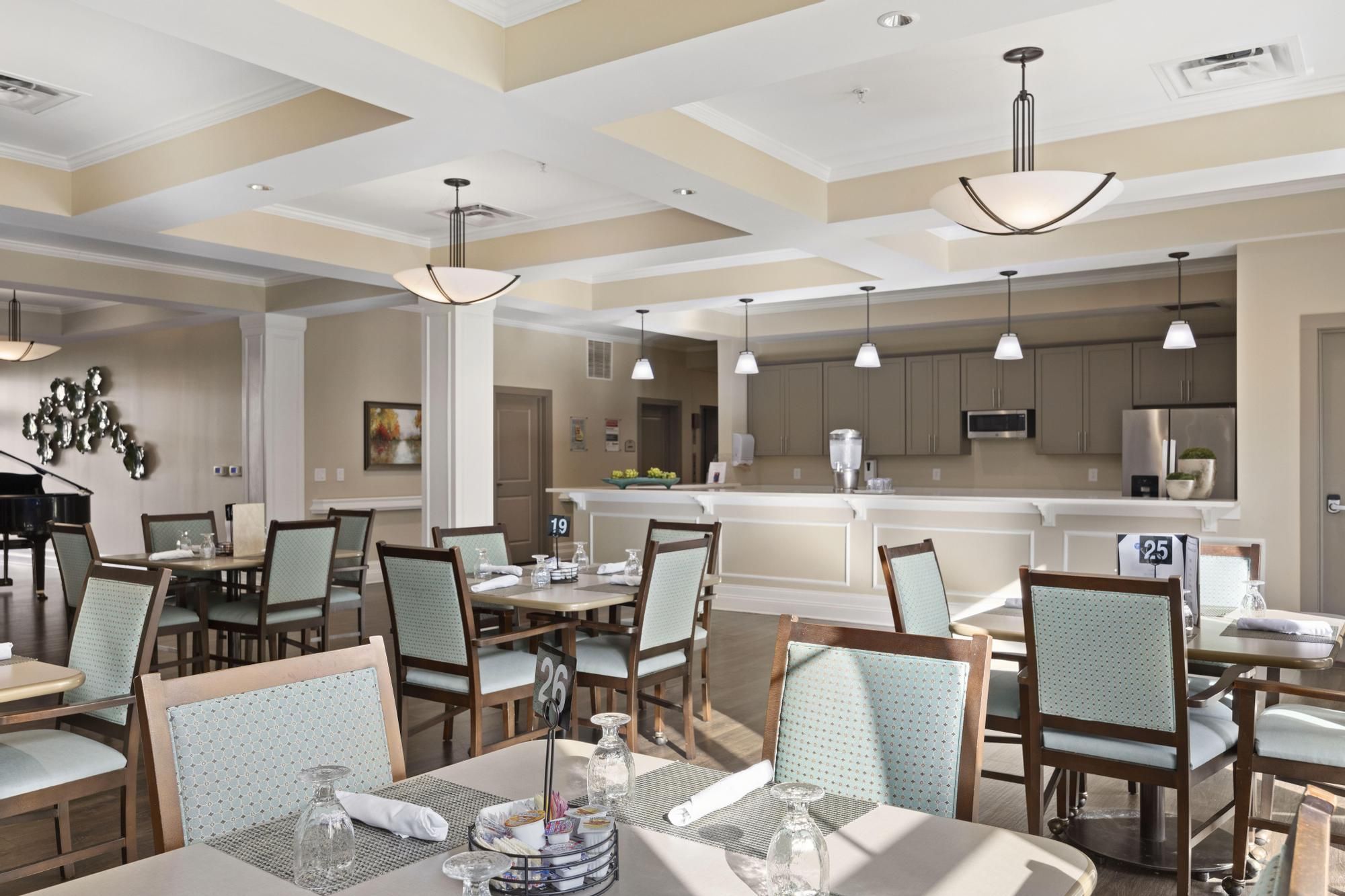 A bright, modern dining area with tables and chairs, ceiling lights, and a view of the kitchen in the background.