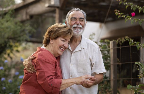 Smiling elderly couple hugging outdoors in a garden, both looking happy and relaxed.
