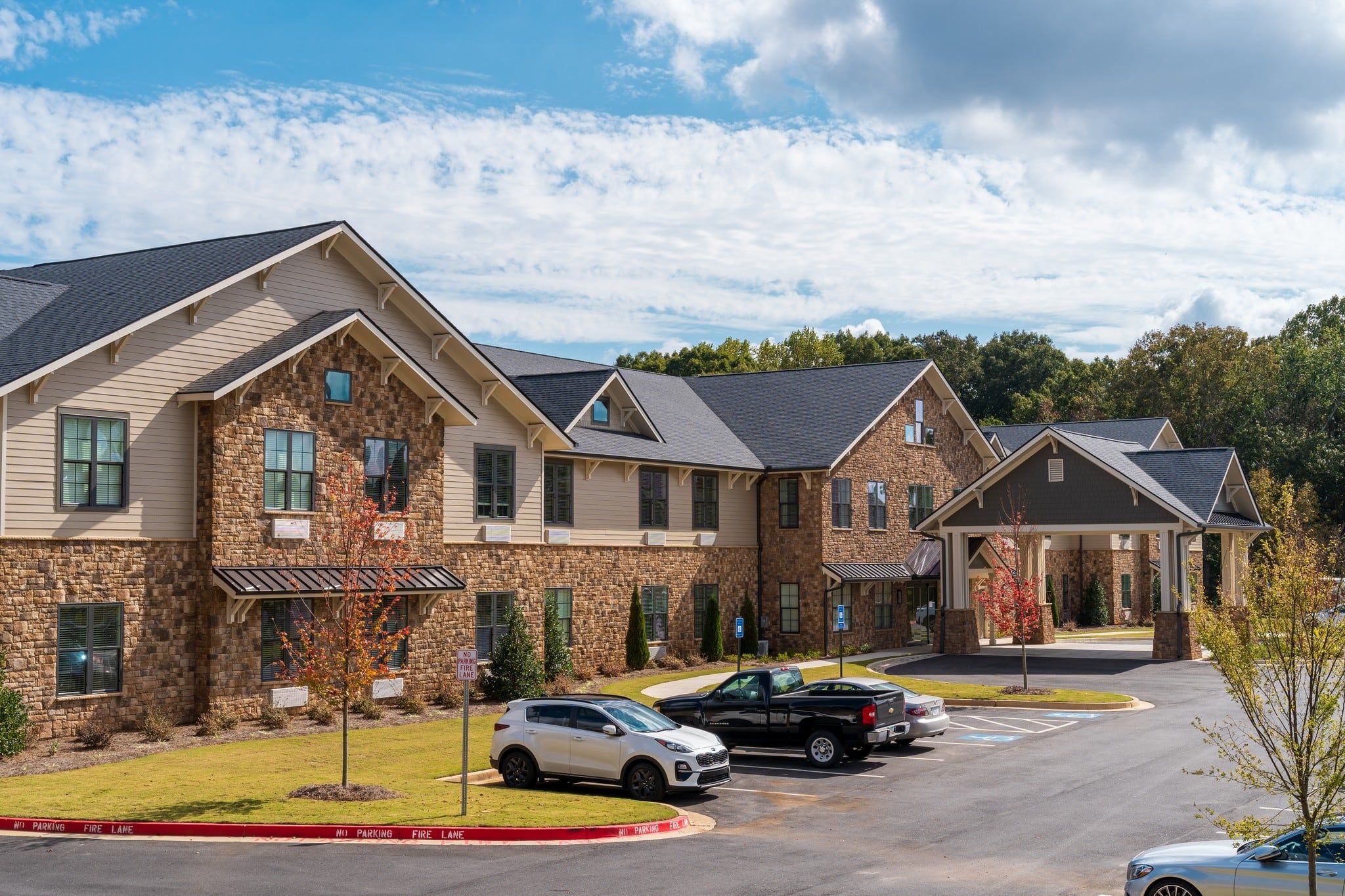 Residential complex with stone facades, multiple peaked roofs, and parked cars under a partly cloudy sky.