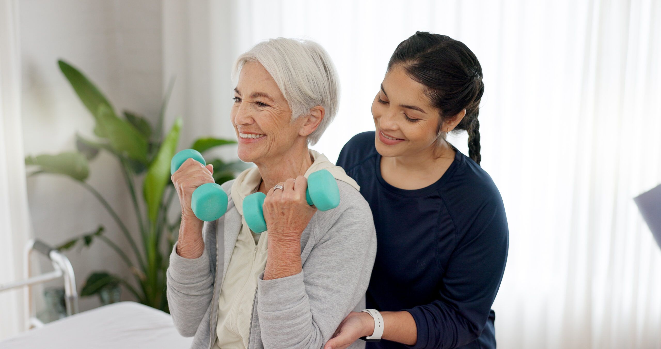 Elderly woman lifts dumbbells with assistance from a caregiver in a bright room.