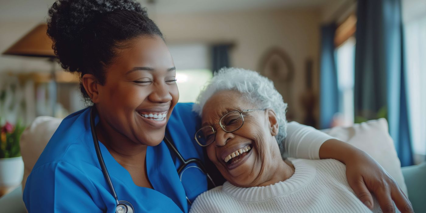 Nurse in blue scrubs with stethoscope hugs and laughs with an elderly woman wearing glasses and a white sweater.