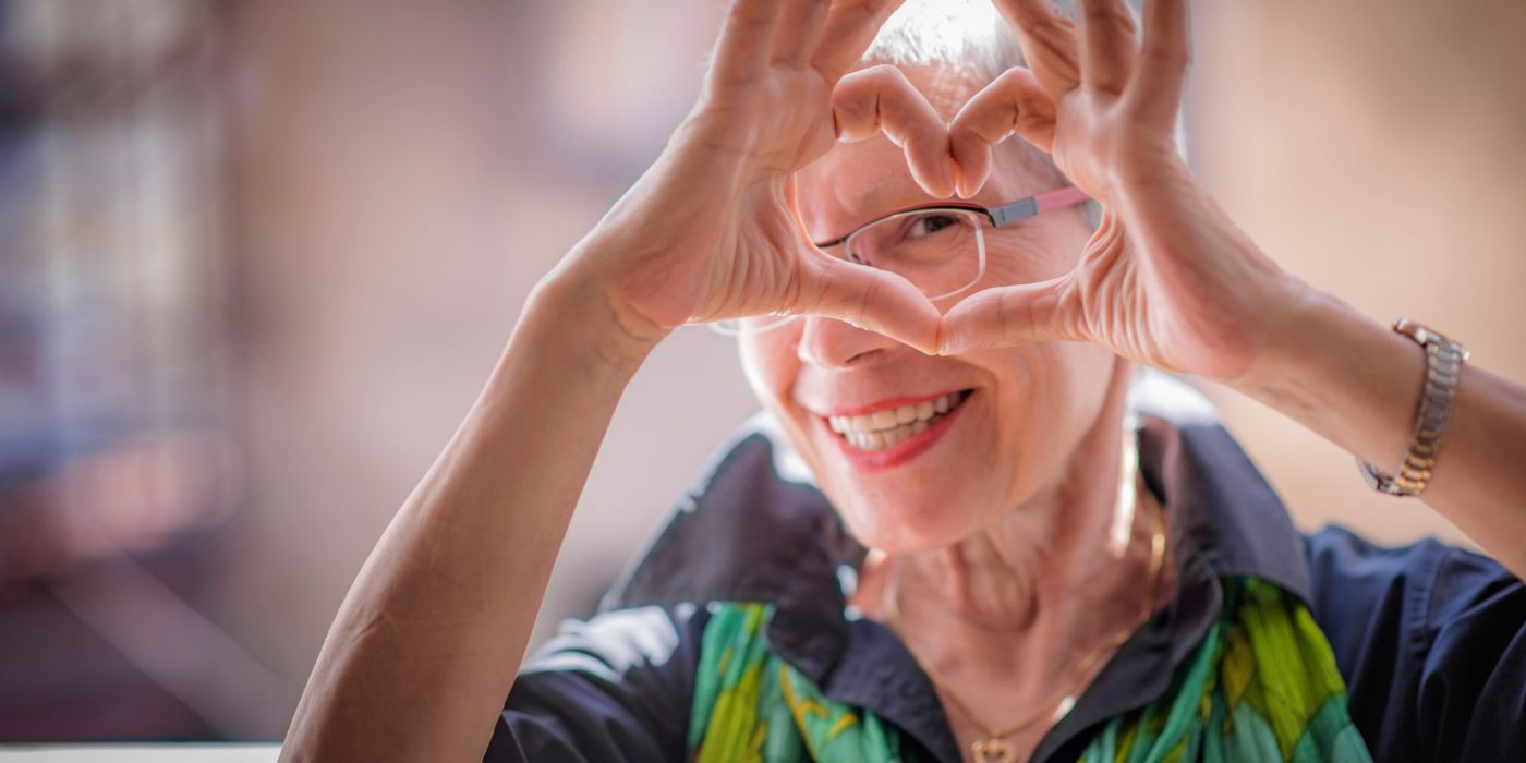 Elderly woman smiling and making a heart shape with her hands over her eyes, wearing glasses and a green scarf.