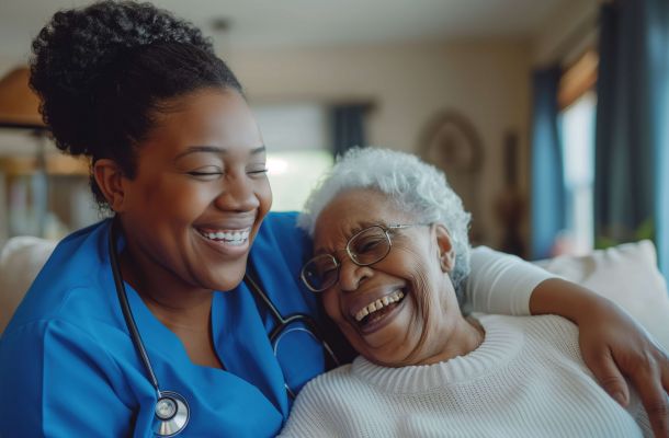 Nurse in blue scrubs with stethoscope hugs and laughs with an elderly woman wearing glasses and a white sweater.