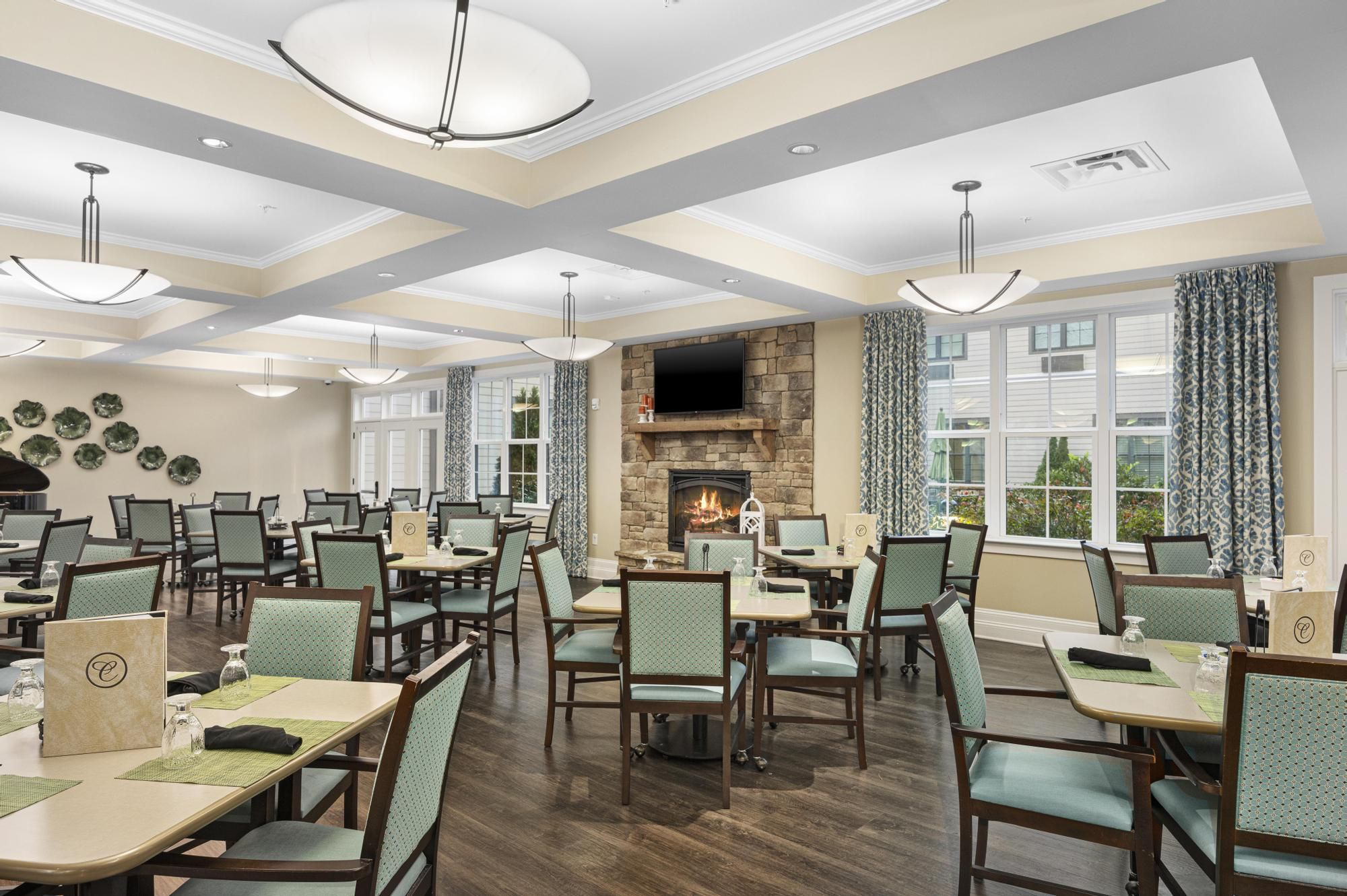 A spacious dining room with green chairs, set tables, and a stone fireplace, lit by large ceiling lights.