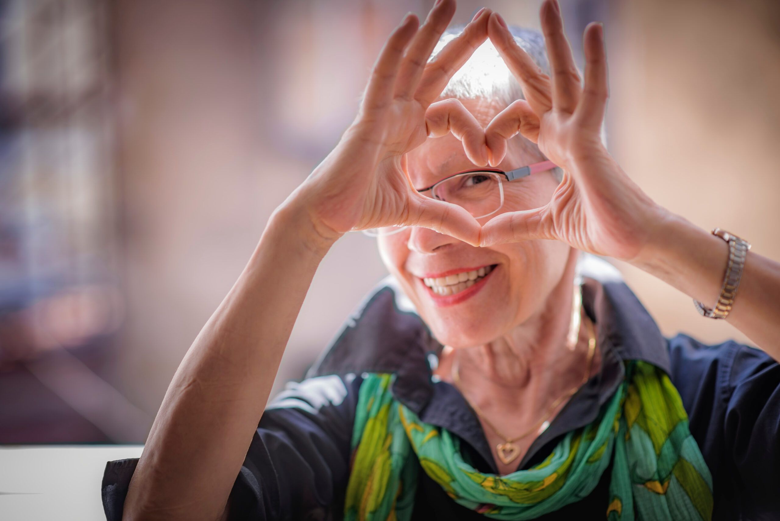 Elderly woman smiling and making a heart shape with her hands over her eyes, wearing glasses and a green scarf.