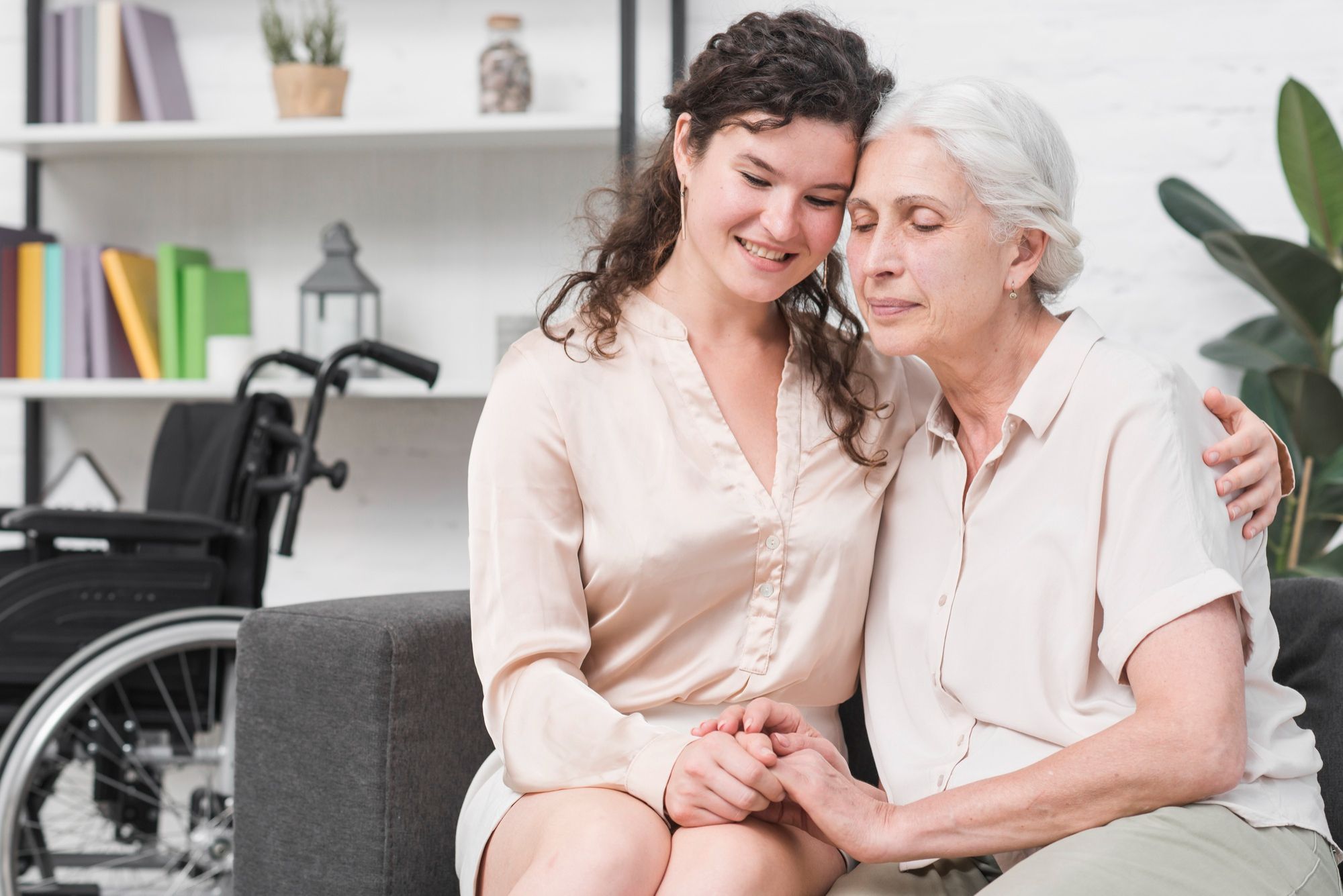 Young woman hugging elderly woman on couch, wheelchair in background, both smiling and holding hands.