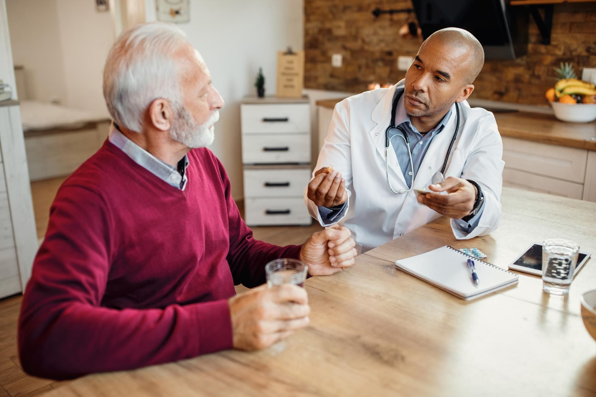 A doctor explains medication to an older man at a kitchen table; both hold glasses of water.