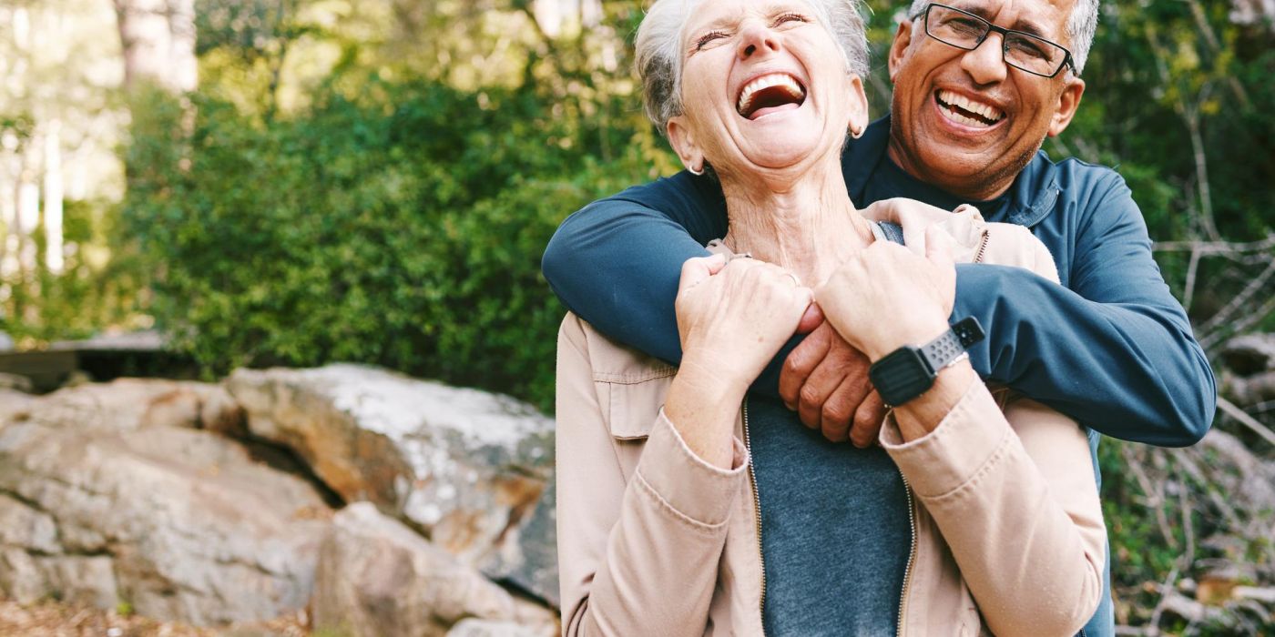 An older couple laughs joyfully as they hug each other outdoors in a wooded park.