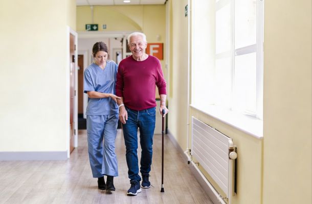 Nurse assists smiling elderly man with a walking stick as they walk down a bright hallway.