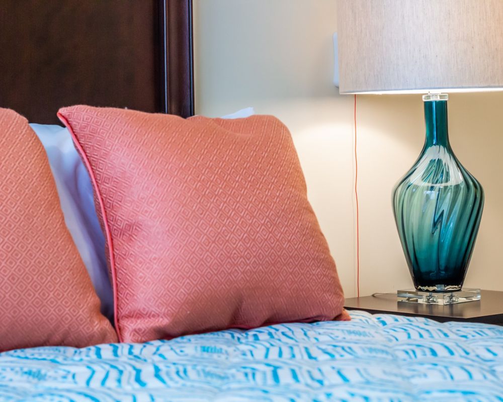 colorful pillows and blankets on a bed next to a pretty lamp and table