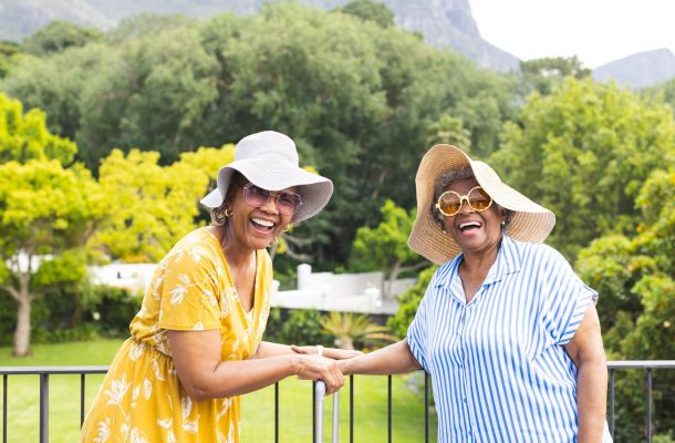 Two older women in sun hats and sunglasses smile and hold hands outdoors with trees in the background.