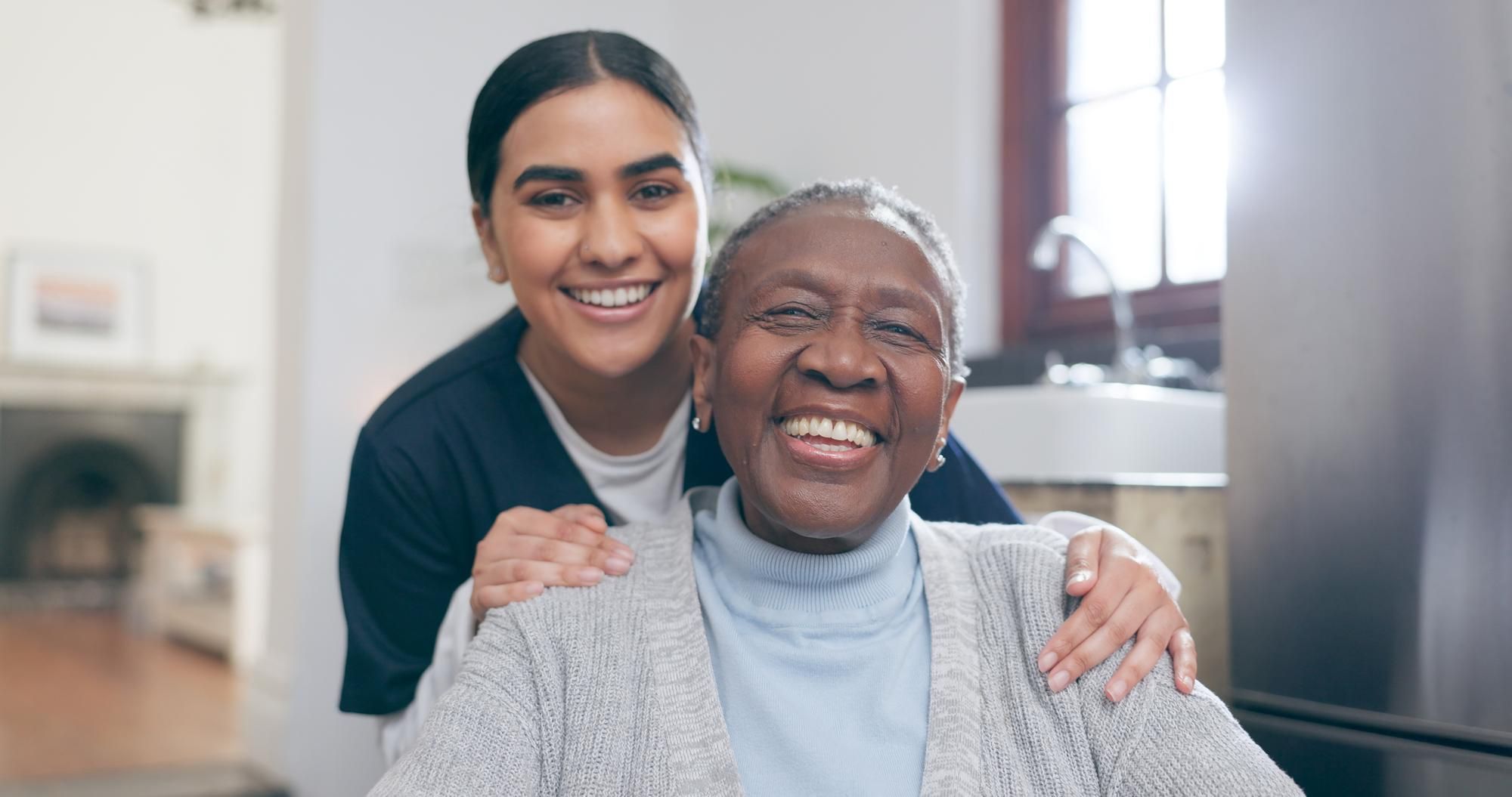 Smiling elderly woman sits while a younger woman stands behind her, both looking happy indoors.