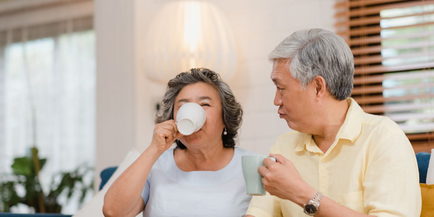 Older couple sitting on a couch at home, enjoying hot drinks and smiling together.