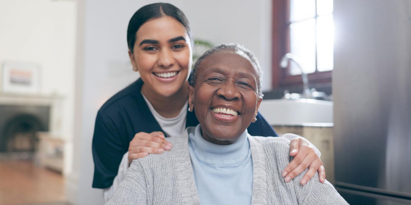 Smiling elderly woman sits while a younger woman stands behind her, both looking happy indoors.