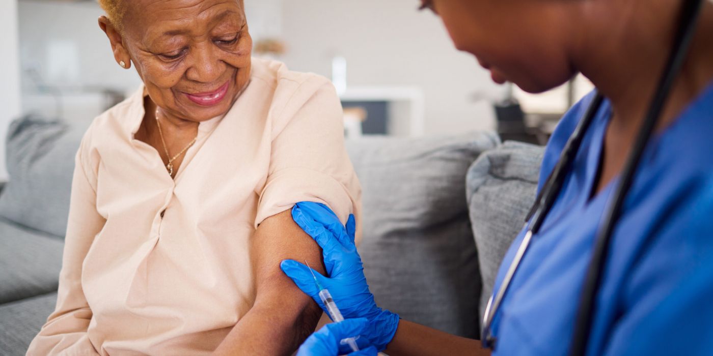 Smiling elderly woman receives a vaccine shot in her arm from a nurse wearing blue gloves.