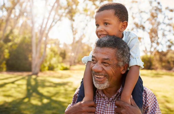 Smiling older man gives a young boy a piggyback ride outdoors on a sunny day in a park.
