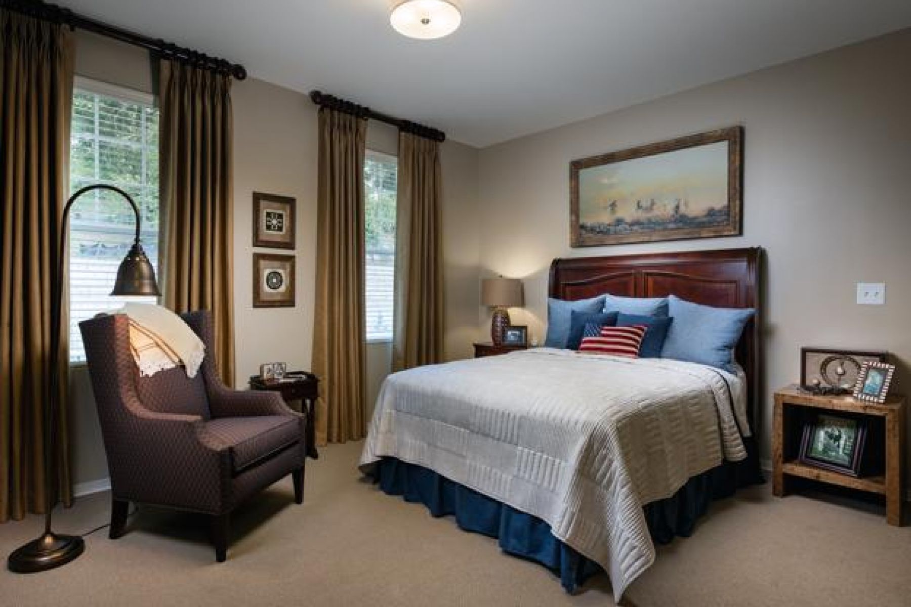  A cozy bedroom with a bed, armchair, lamp, and framed artwork on beige walls and brown curtains.Camellia Place
