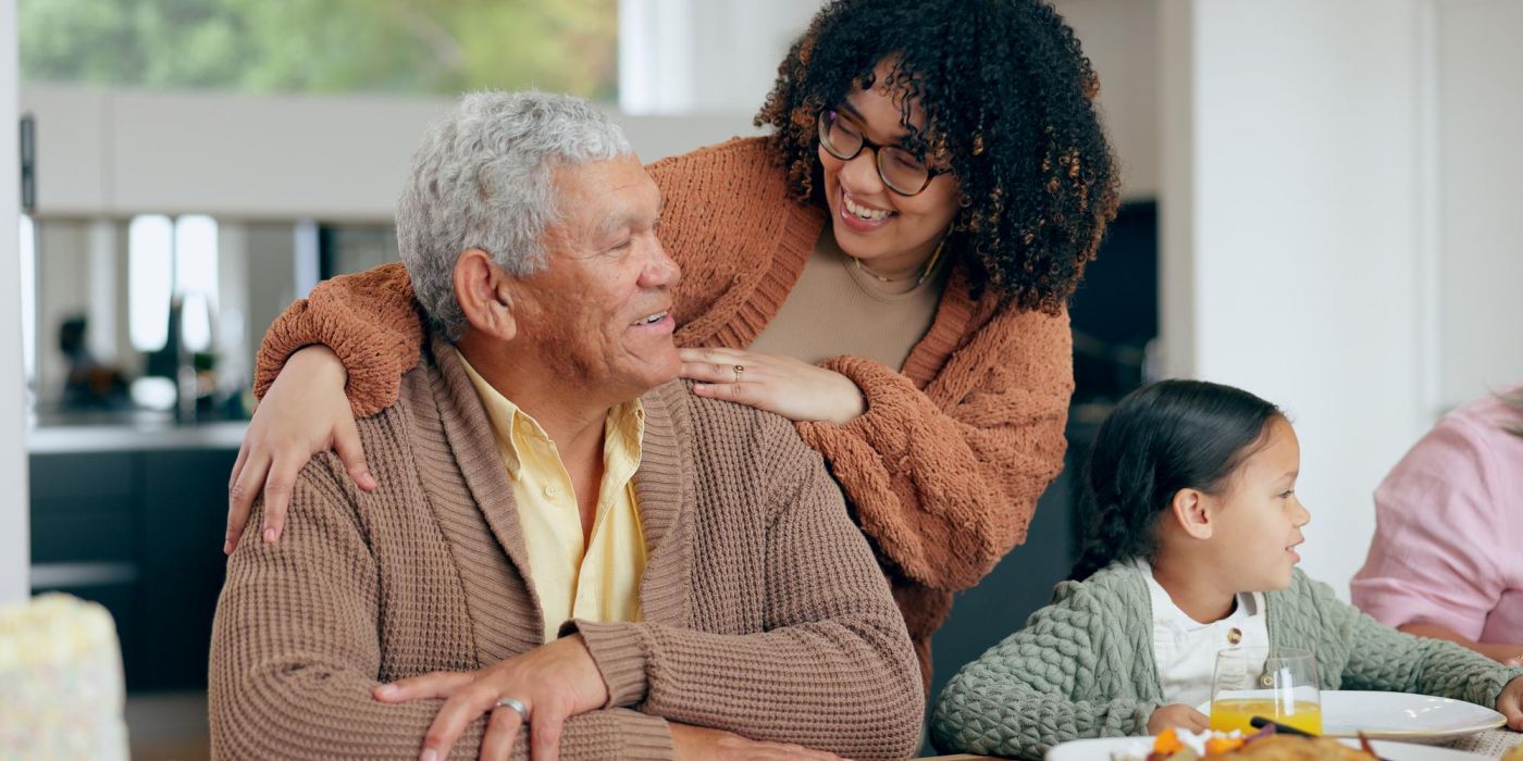 A Family Visit brings joy as an elderly man smiles at a woman behind him, while a young girl sits nearby at the dining table.