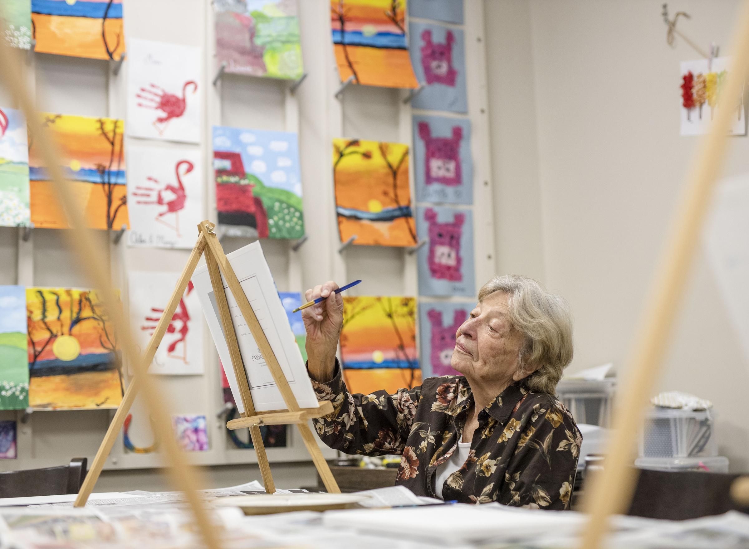 Camellia Place Camellia Place An elderly woman paints at an easel in a room decorated with colorful artwork and paintings on the wall.