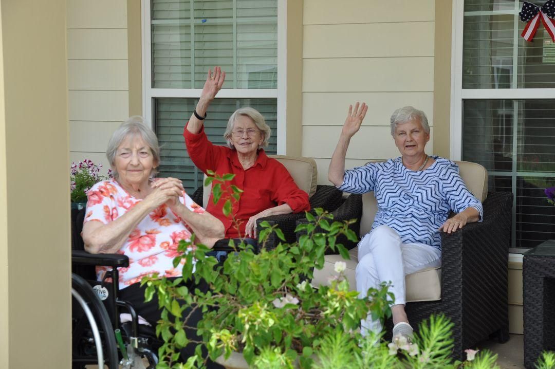Camellia Place Three elderly women sitting outdoors, two of them waving and smiling, with plants in the foreground.