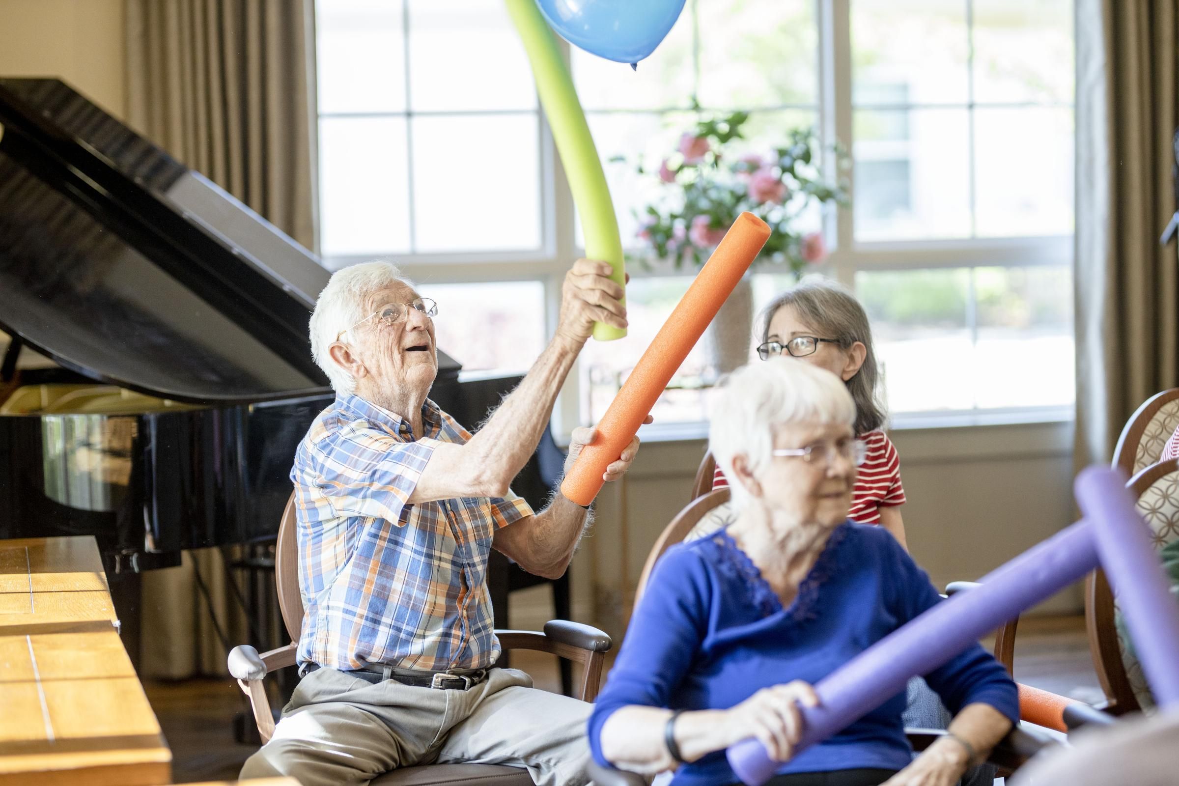 Camellia Place Seniors play with colorful foam noodles and balloons while seated indoors near a piano.