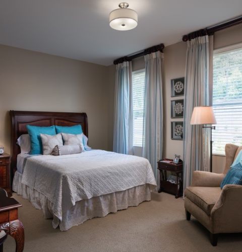 Camellia Place Cozy bedroom with a neatly made bed, armchair, lamps, curtains, and soft natural light from windows.