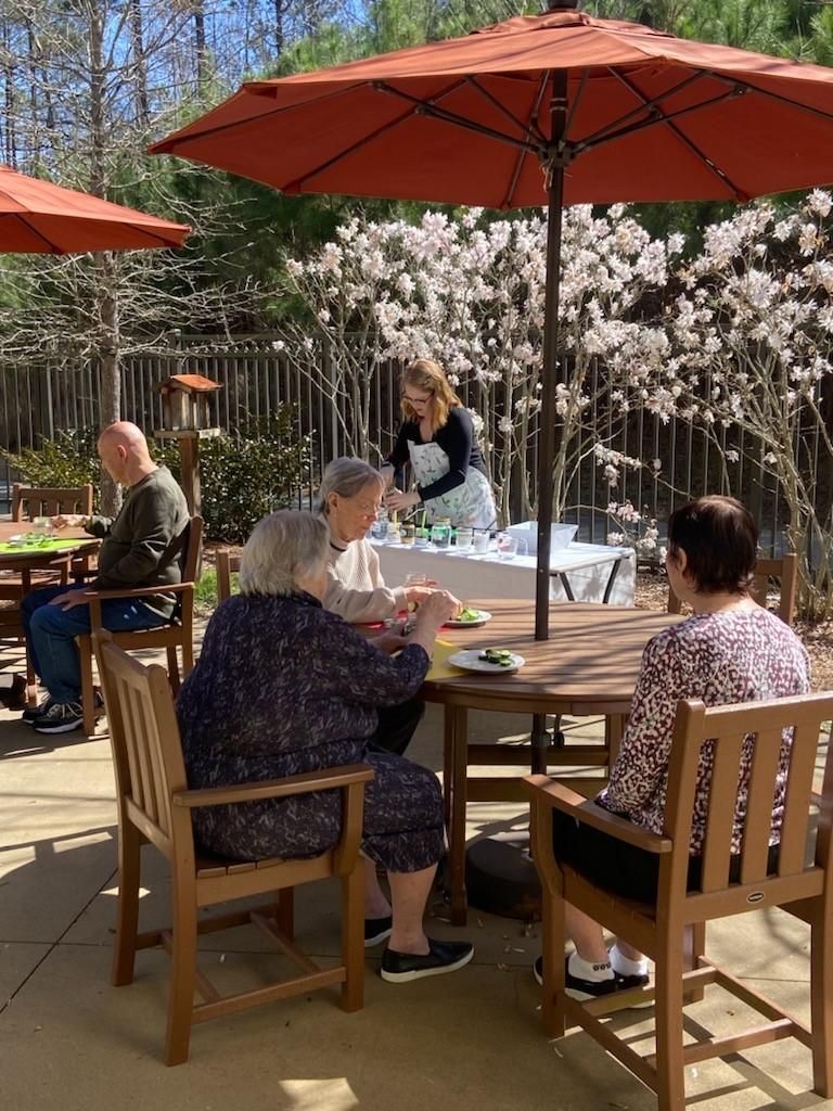 Camellia Place Four older adults sit at patio tables under umbrellas; a woman stands at a serving table in the background.