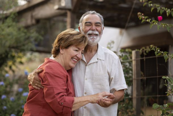 Smiling elderly couple hugging outdoors in a garden, both looking happy and relaxed.
