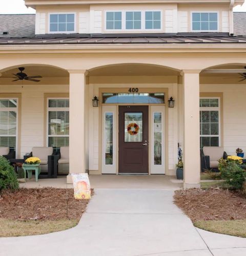 Camellia Place Single-story house entrance with a covered porch, columns, patio chairs, and a wreath on the front door.