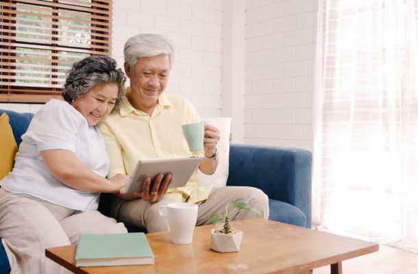 Smiling older couple sits on a couch, looking at a tablet together, with coffee mugs on a wooden table.