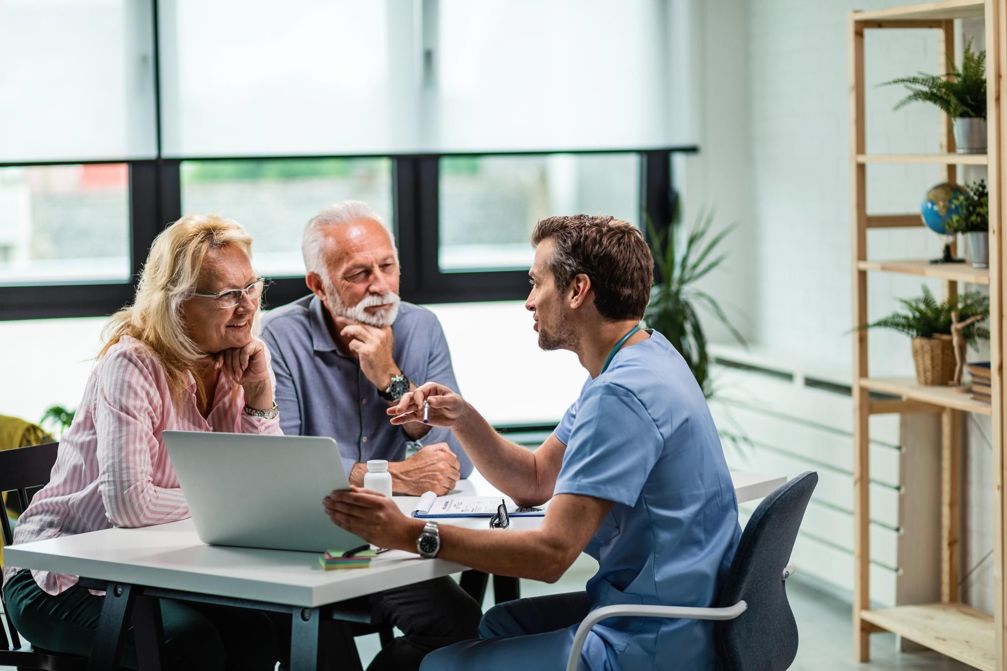 A doctor discusses something with an older couple, using a laptop and documents at a table in an office.