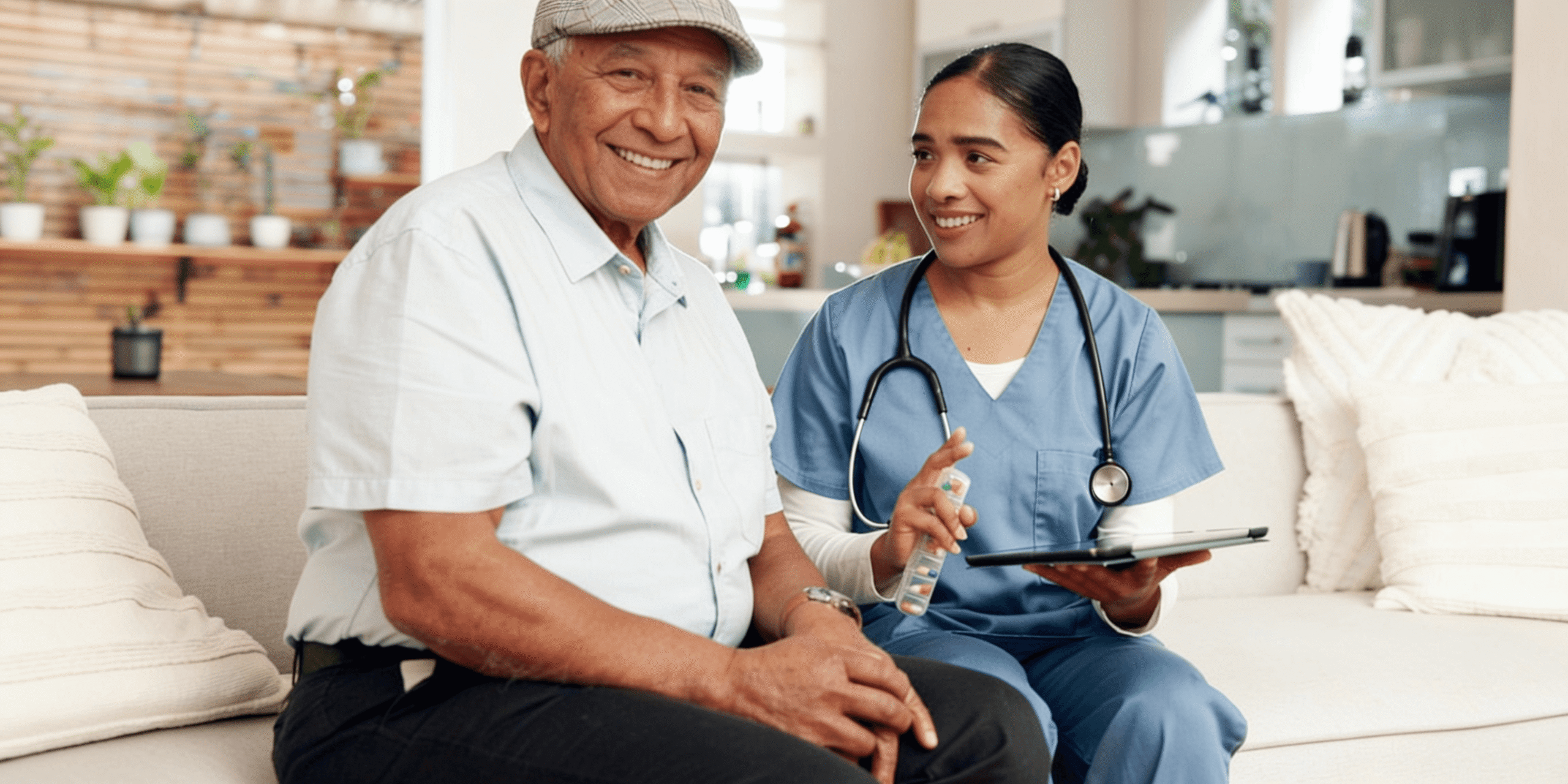 An elderly man smiles while sitting next to a nurse with a stethoscope and tablet in a bright living room.