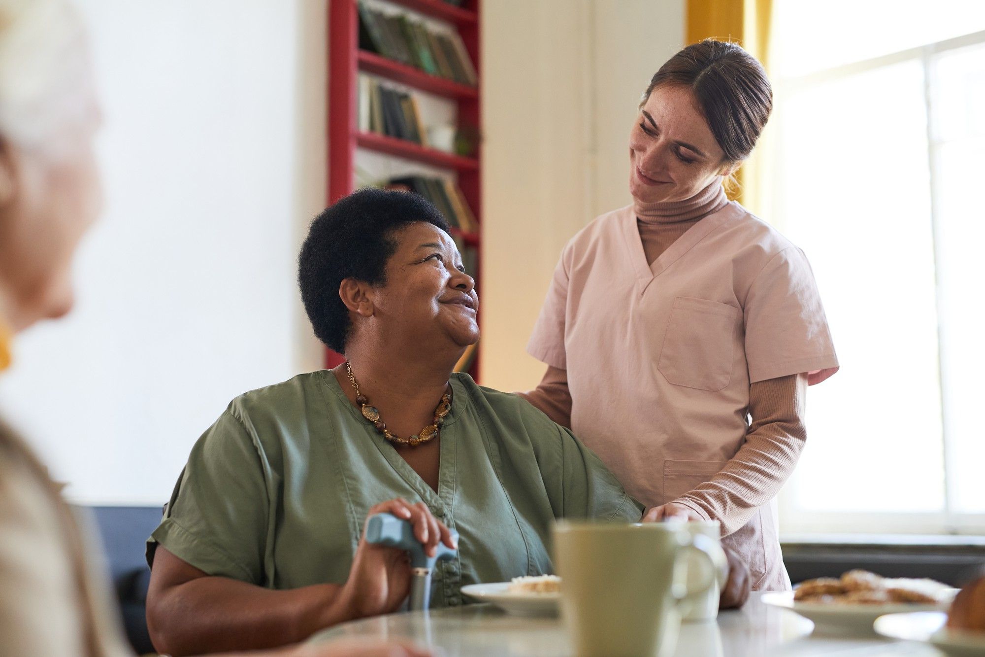 Smiling caregiver stands beside a seated woman, both sharing a warm interaction in a cozy room.