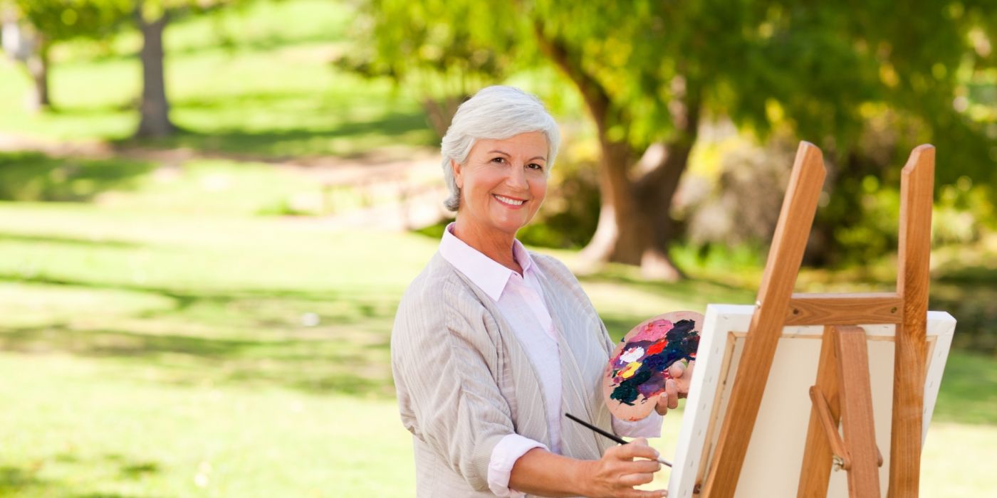 Smiling older woman painting at an easel outdoors in a park, holding a palette with colorful paint.