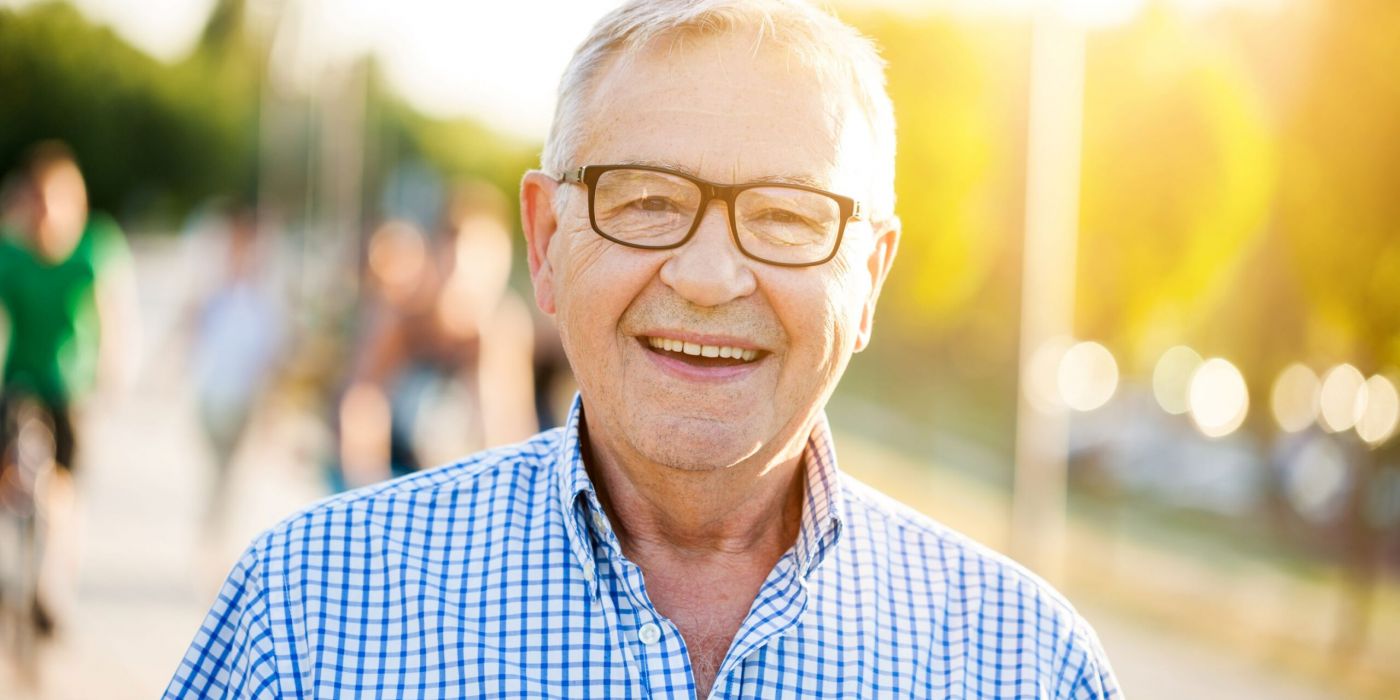 Smiling older man wearing glasses and a checked shirt, standing outdoors in sunlight.