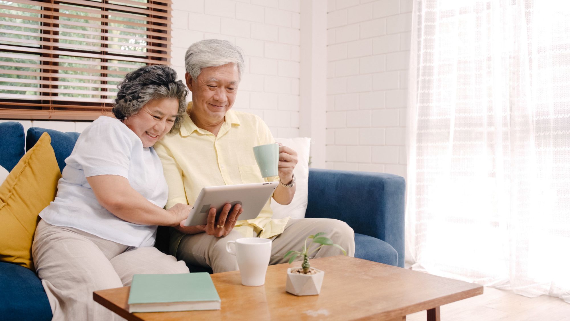 Smiling older couple sits on a couch, looking at a tablet together, with coffee mugs on a wooden table.