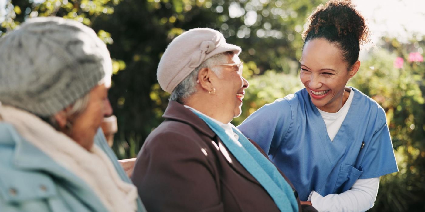 A nurse smiles and talks with two elderly women sitting outside on a bench in a garden.