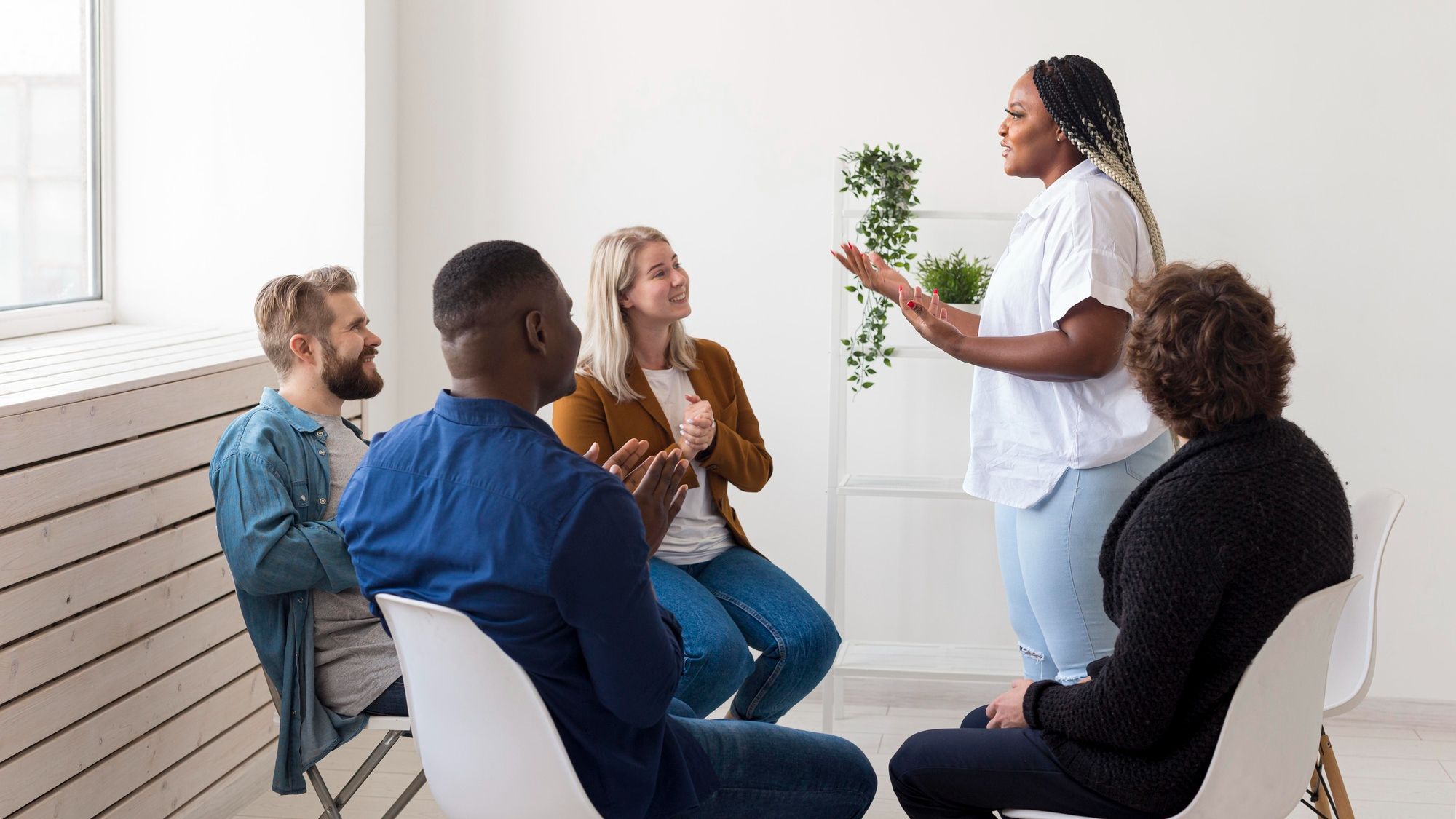 A woman stands speaking to a small group of people sitting in chairs, who are listening and clapping.