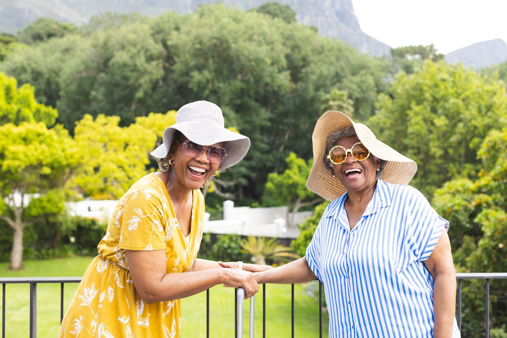 Two older women in sun hats and sunglasses smile and hold hands outdoors with trees in the background.