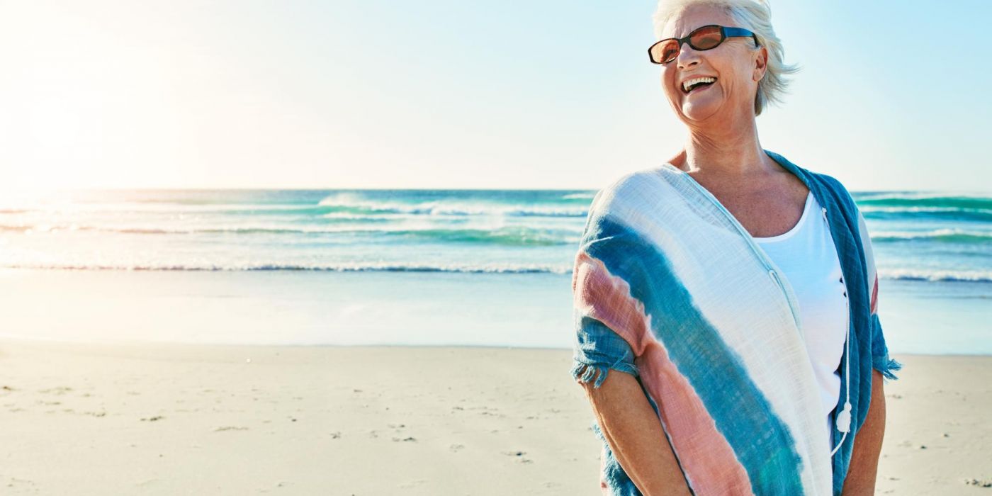 Smiling older woman in sunglasses stands on a sunny beach with waves in the background.