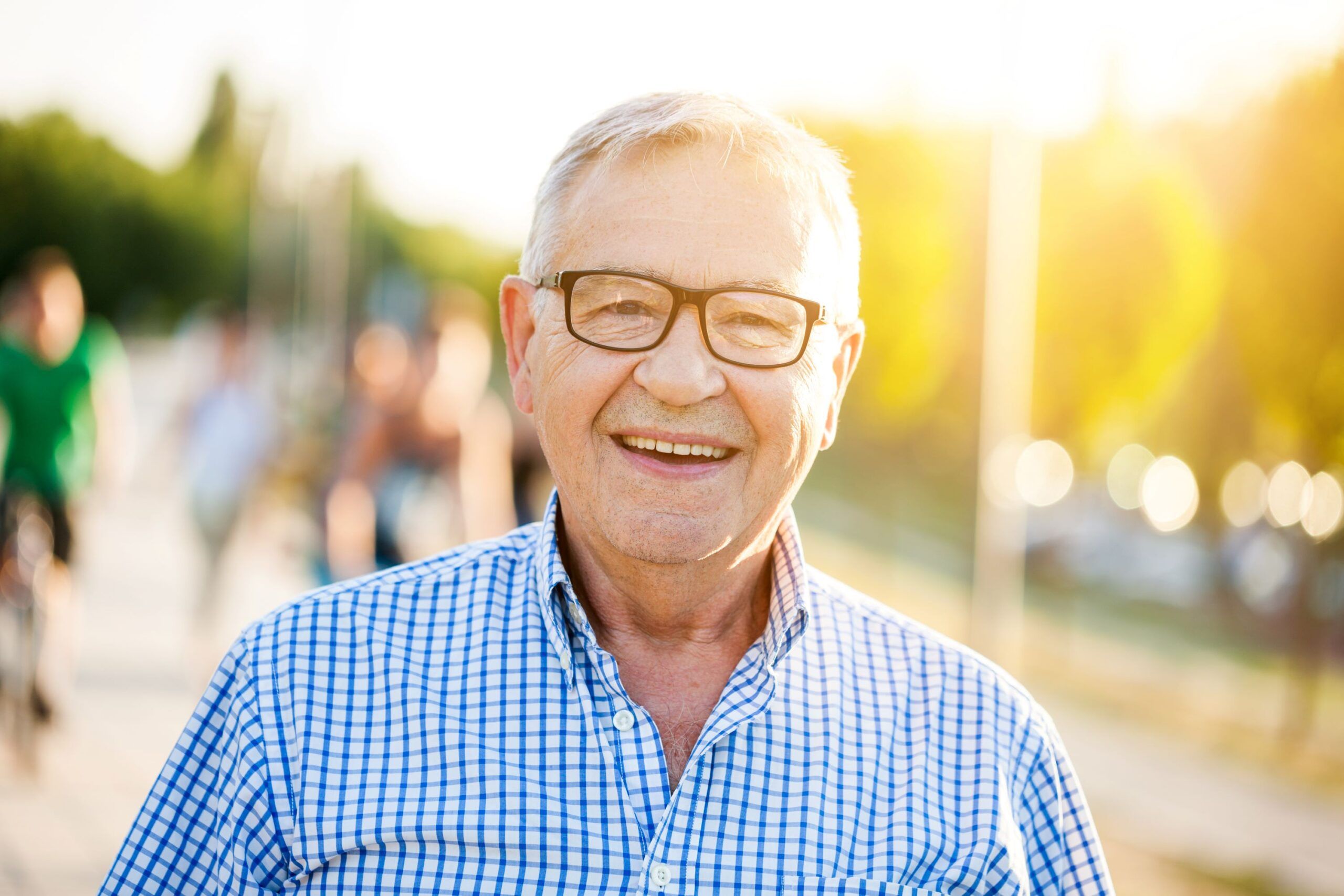 Smiling older man wearing glasses and a checked shirt, standing outdoors in sunlight.